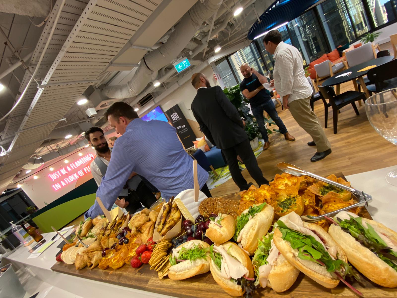 A corporate catering lunch board in the foreground of a seminar. There are people standing in the background chatting