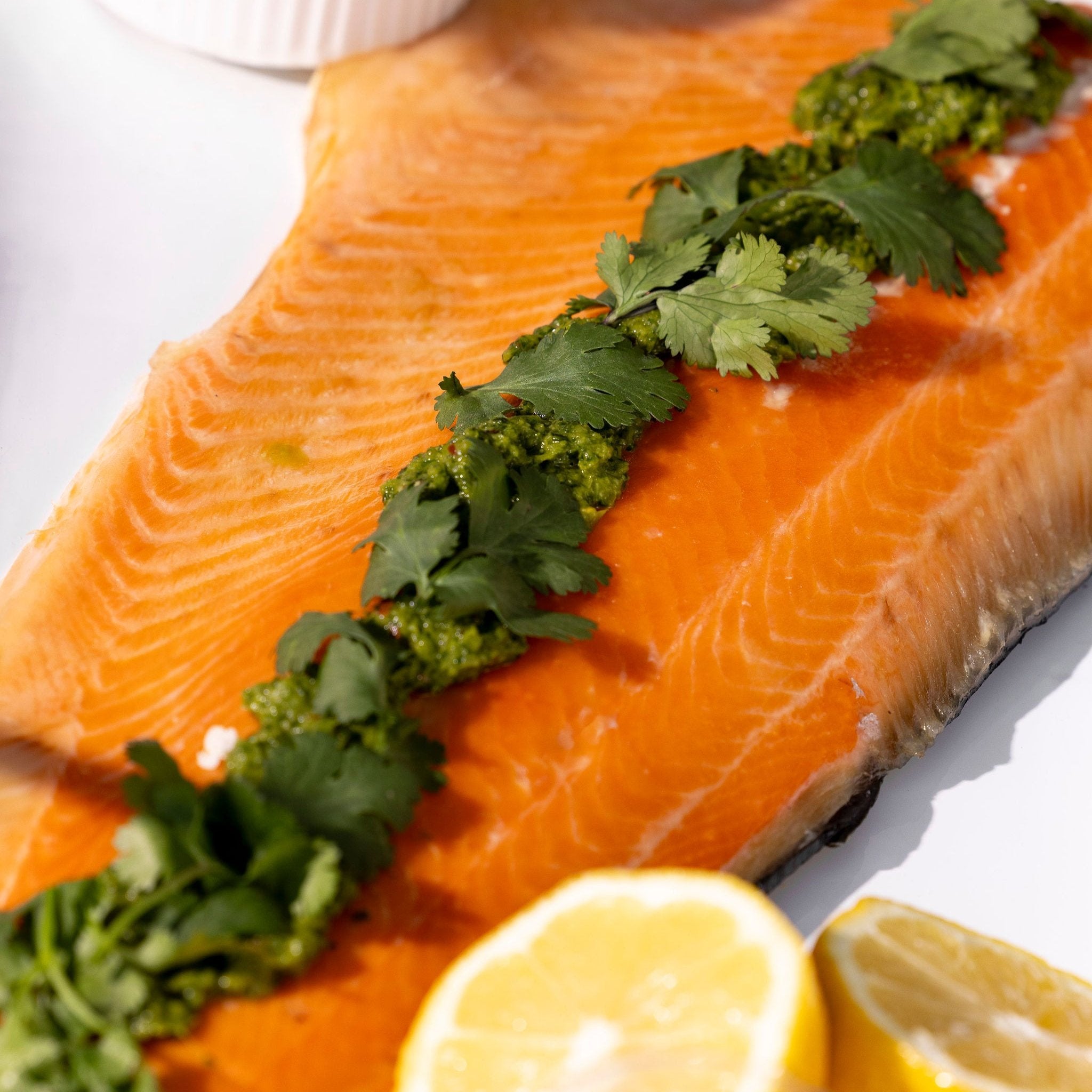 A side of salmon, dressed with chimichurri & coriander leaves sits on a white platter.